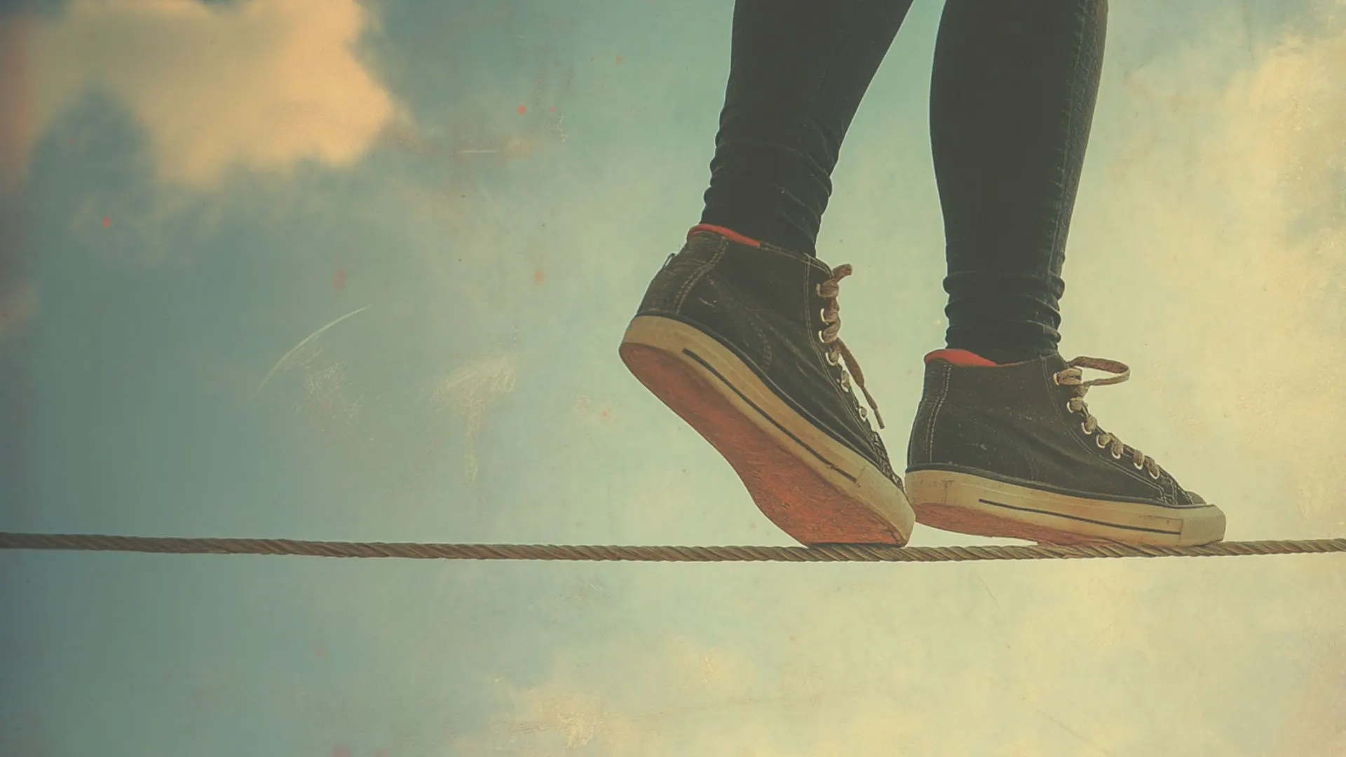 Person wearing dark sneakers standing on a tightrope against a cloudy sky