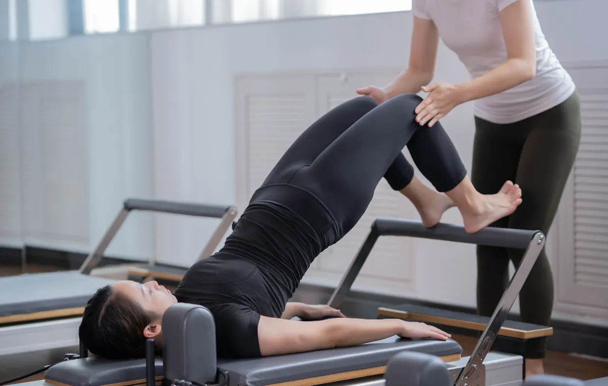 A Pilates instructor gently guides a woman's legs as she lifts into a bridge on a reformer.