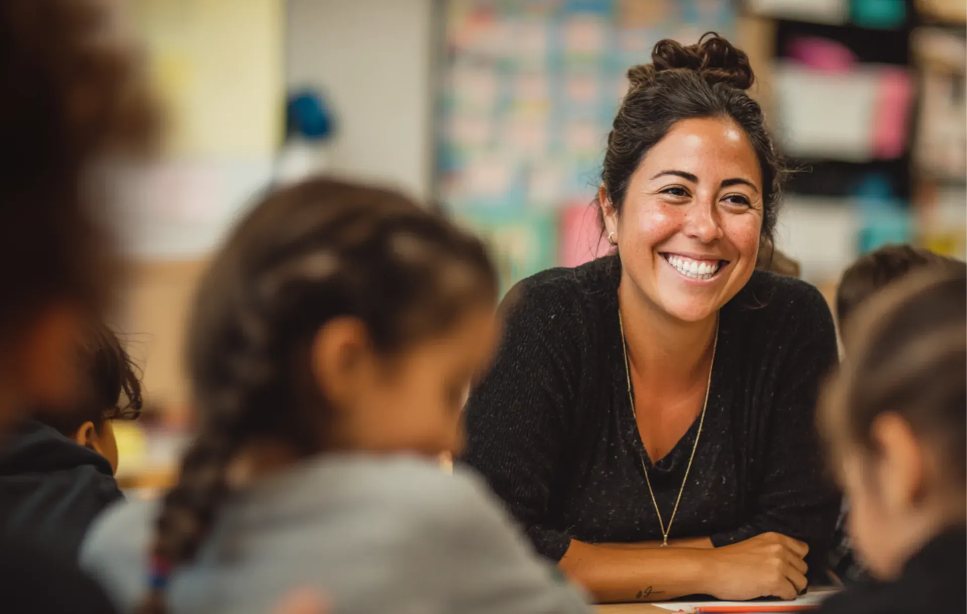 A teacher smiles warmly while engaging with students in a colorful classroom