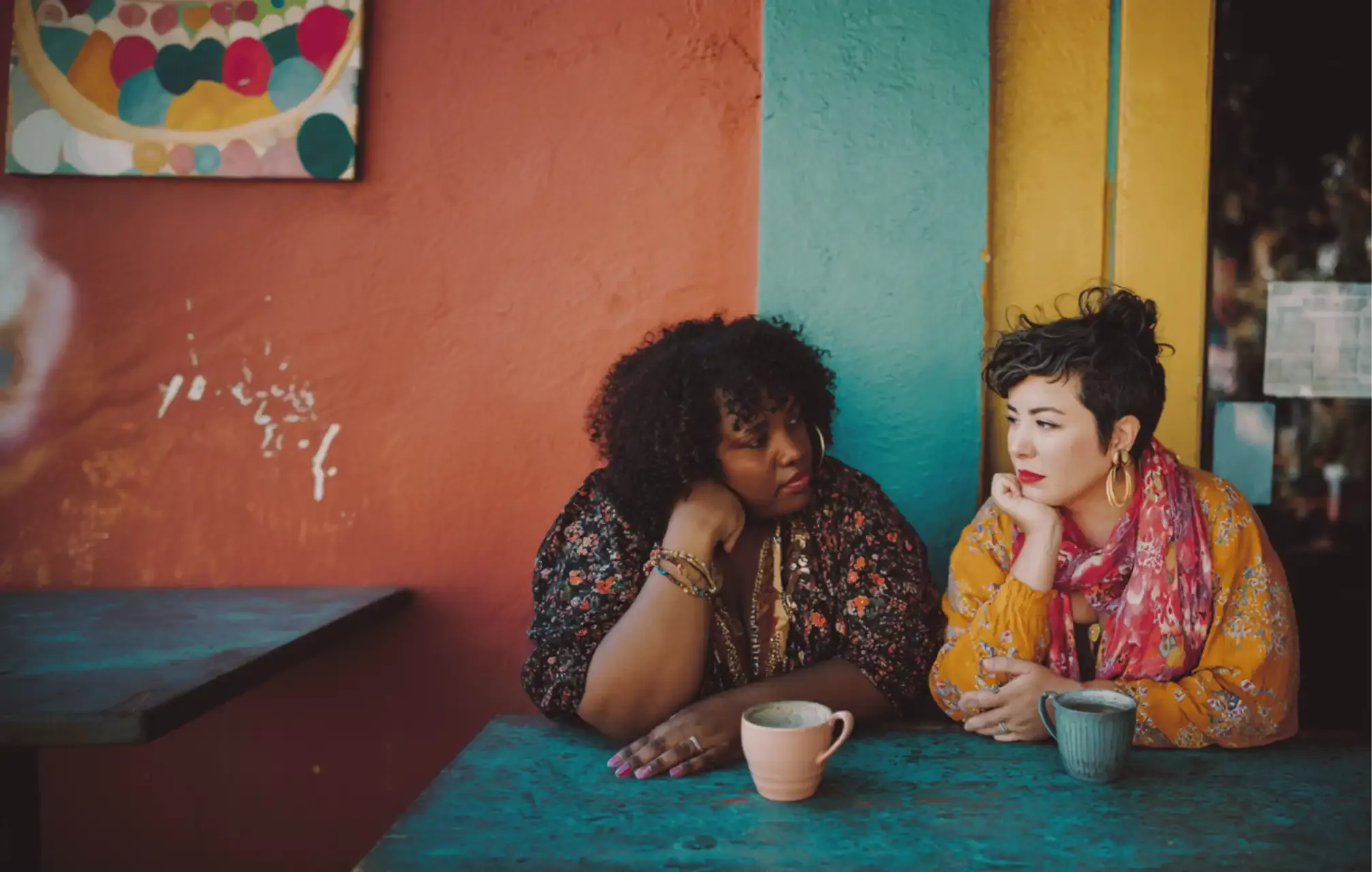 Two women of color sit together at a café table, one listening closely as the other speaks.