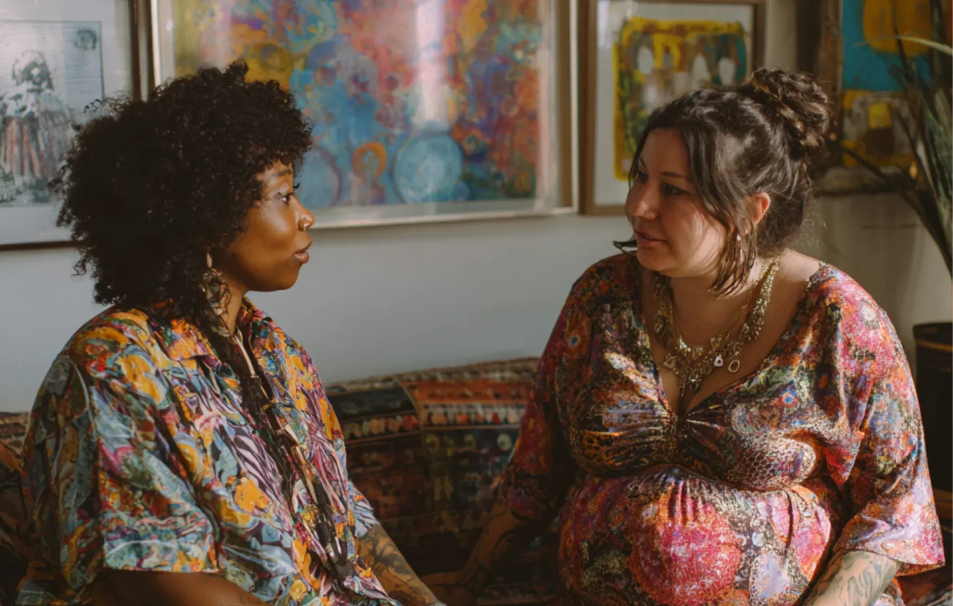 Two women talk closely together in a room surrounded by colorful artwork, one turned attentively.