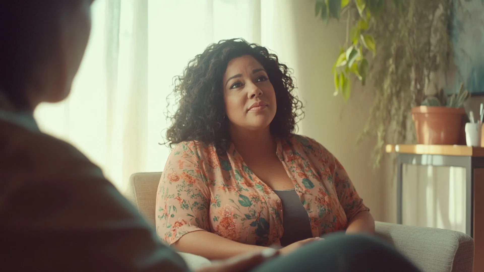 A woman with curly dark hair sits at a table across from another person, looking upward thoughtfully in a bright room with plants and windows