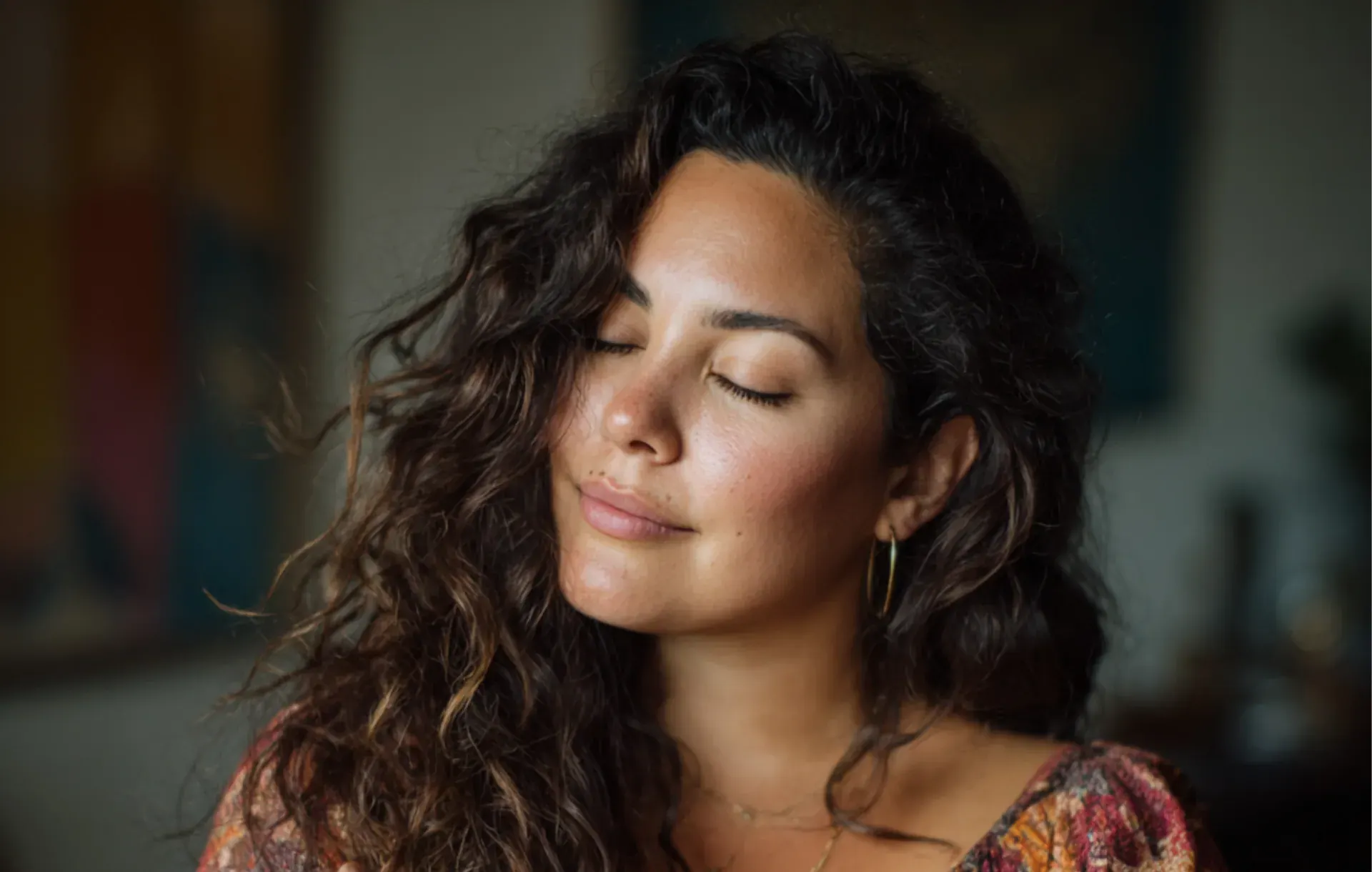 A woman with dark curly hair closes her eyes in soft, warm lighting.