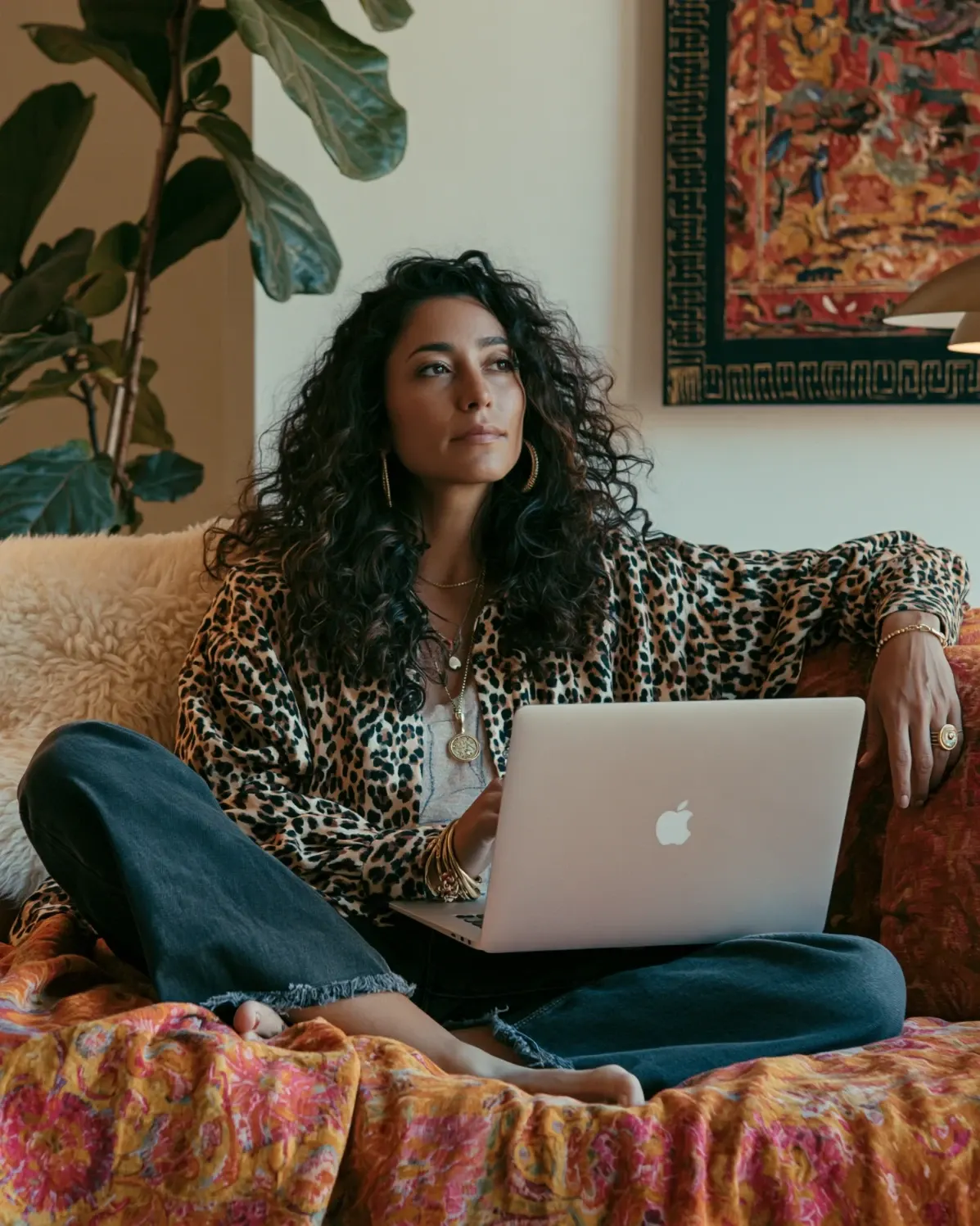 A woman with long curly hair sits cross-legged on a sofa with a laptop, looking up thoughtfully, in a warm living room