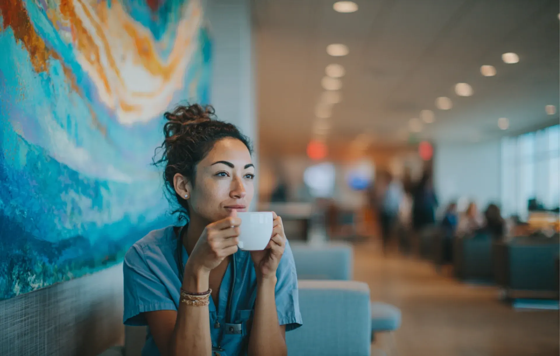 A woman in scrubs pauses for coffee in front of a colorful mural, lost in thought.