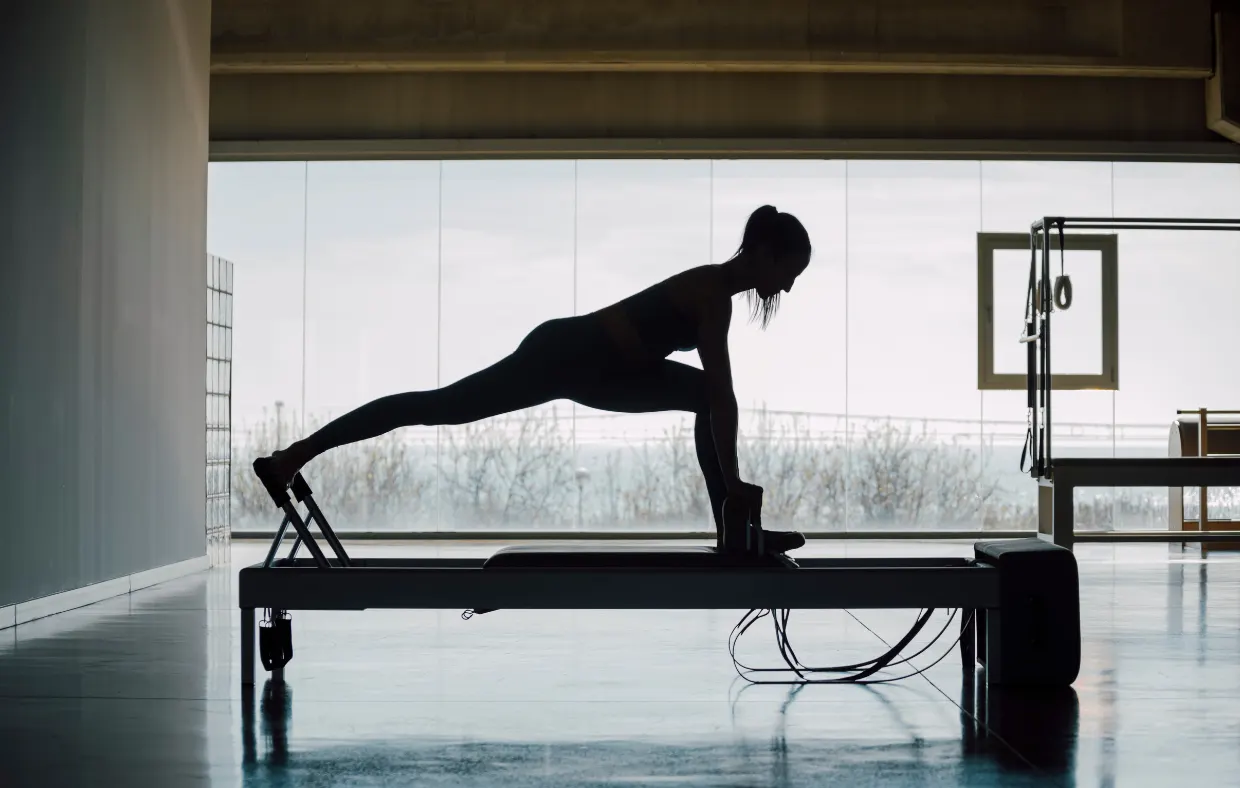 A woman in silhouette stretches into a lunge on a reformer in front of tall windows.