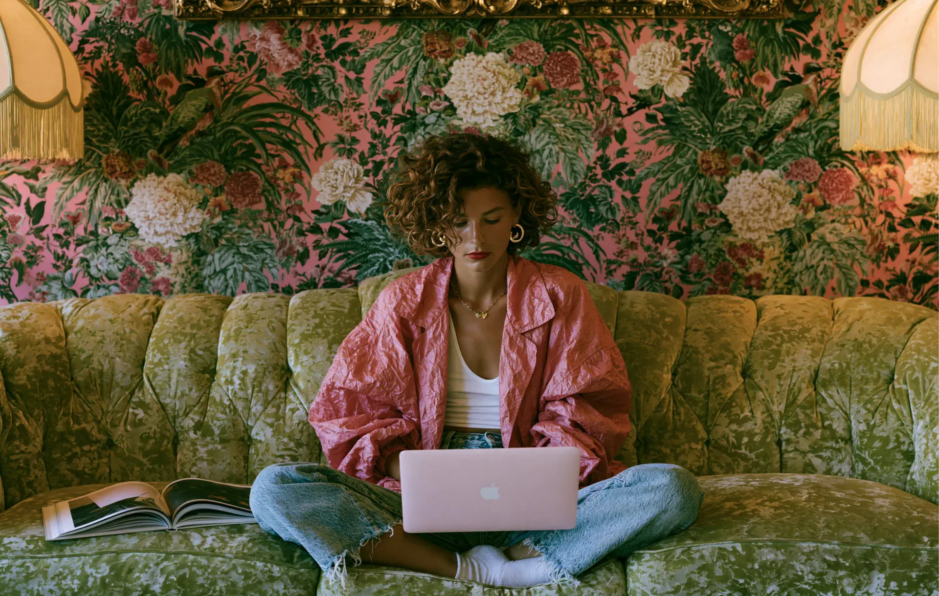 A woman sits cross-legged with a laptop on a green velvet couch in a floral-papered room.