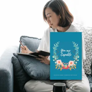 Woman sitting on a gray couch reading an open blue book with floral wreath design