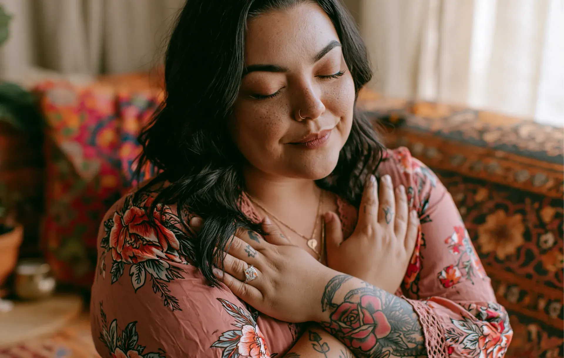 A woman with tattoos in a pink floral top wraps her arms around herself, eyes closed.