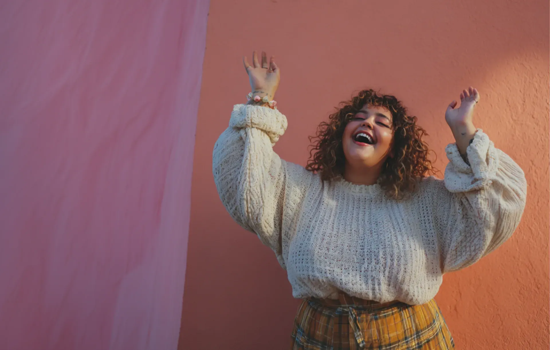A woman throws her arms up dancing joyfully against a pink wall