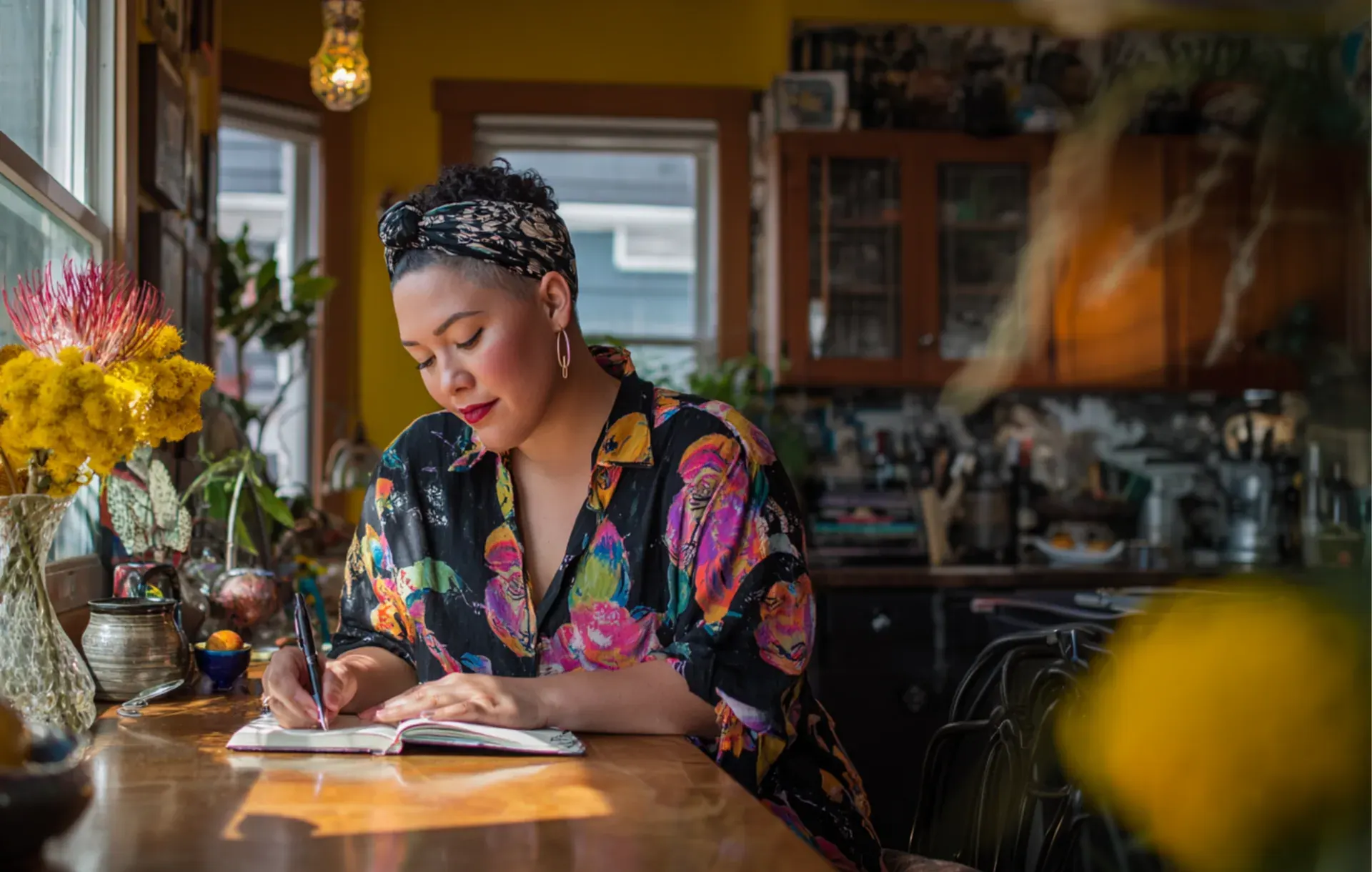 A woman writes in a journal at a colorful kitchen table wearing a headscarf