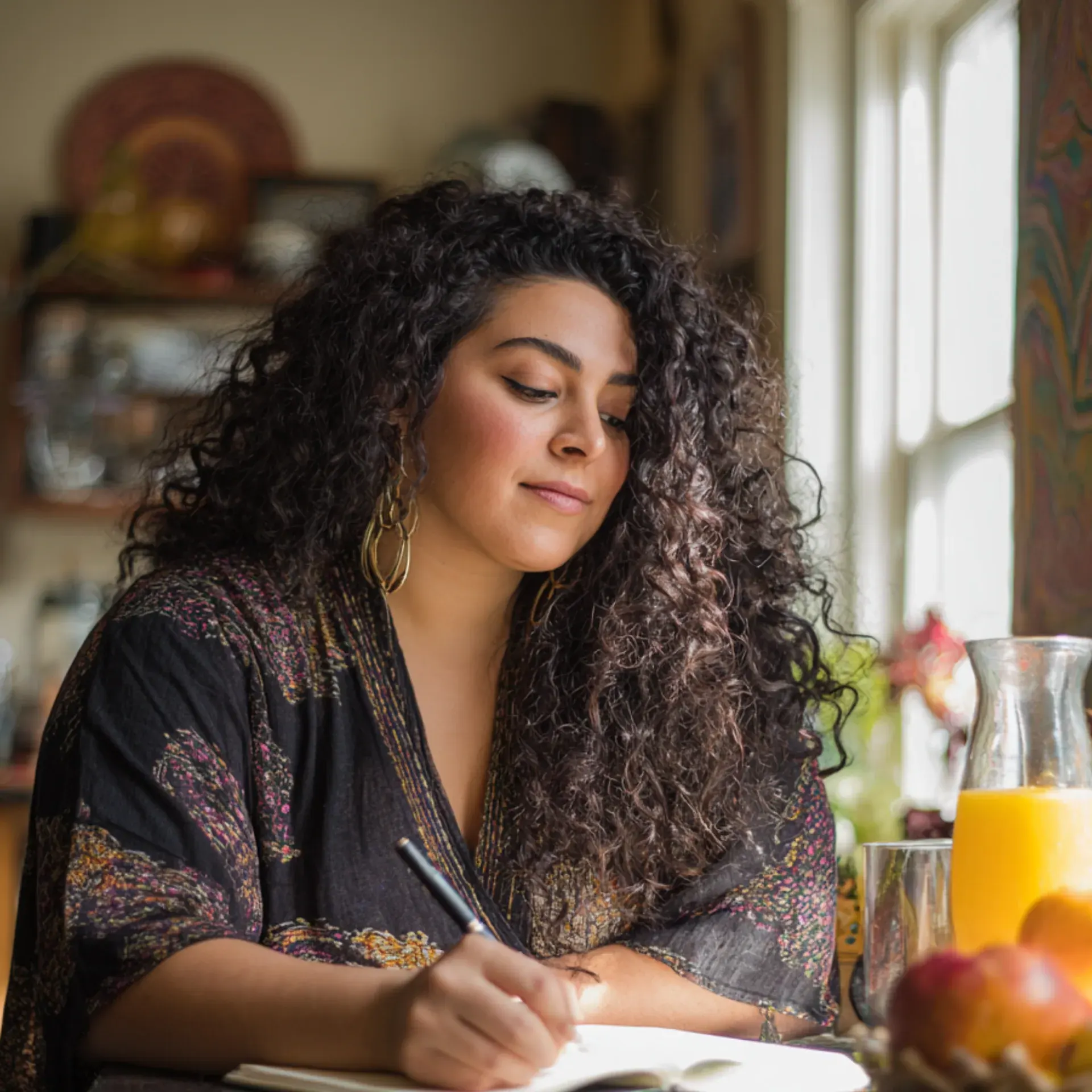 A woman writes in a journal by a sunlit window with orange juice and fruit nearby.