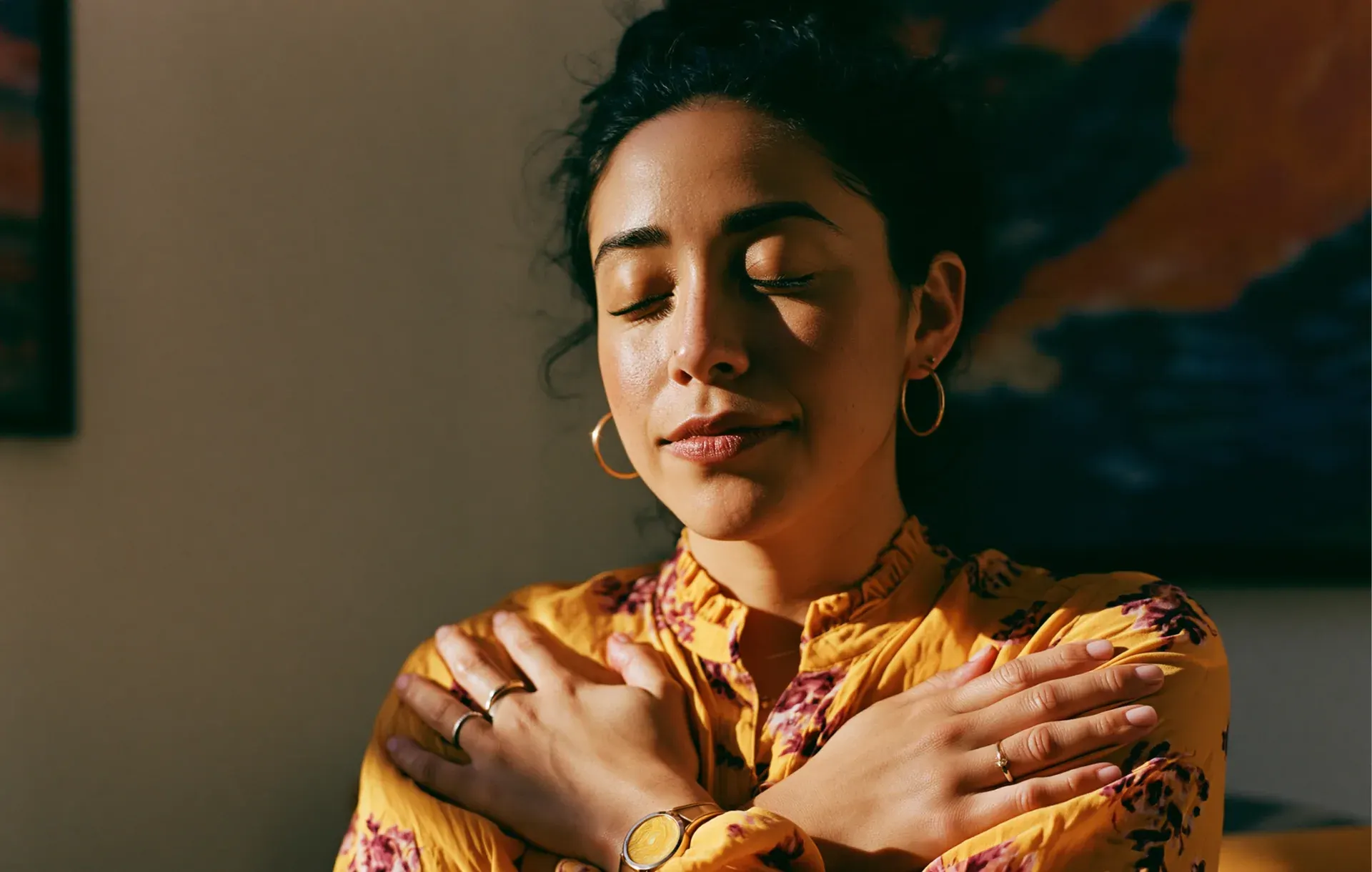 A woman in a yellow floral top closes her eyes and hugs herself gently.
