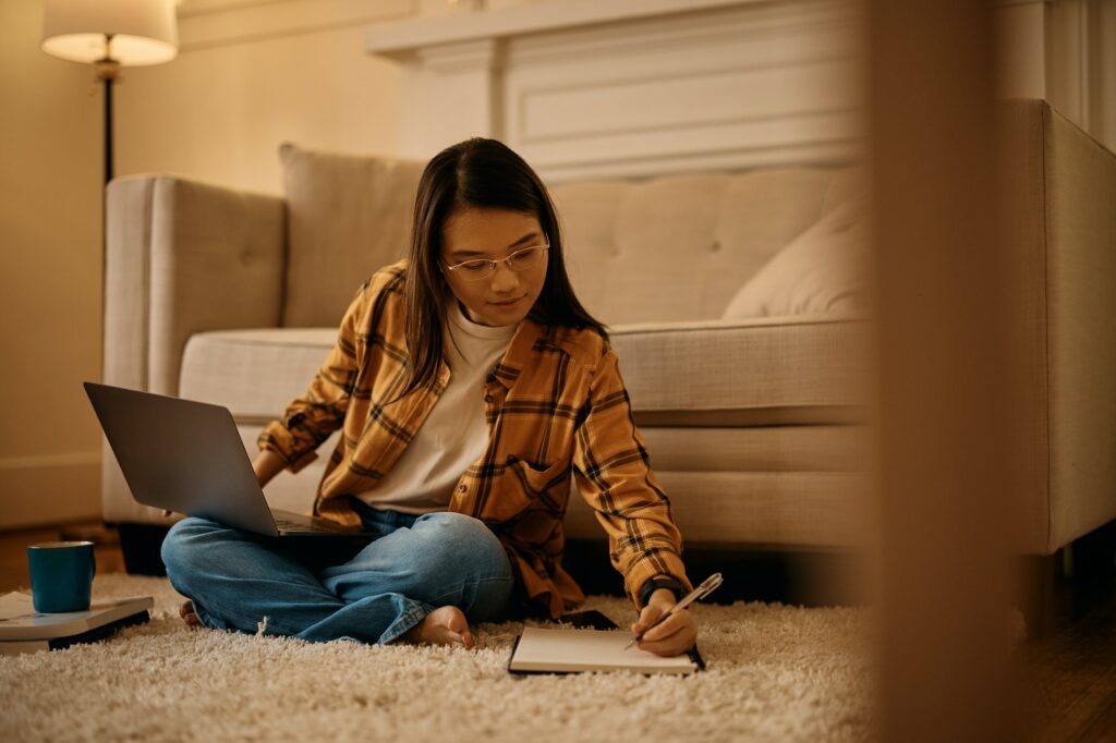 Asian female student writing notes while learning on laptop at home.